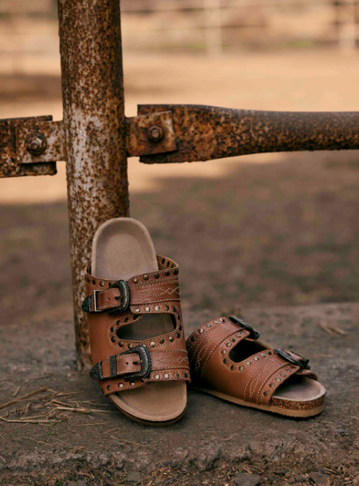 Great Sonora Sunset Sandals in tan with Western buckles and brass studs on rustic background.