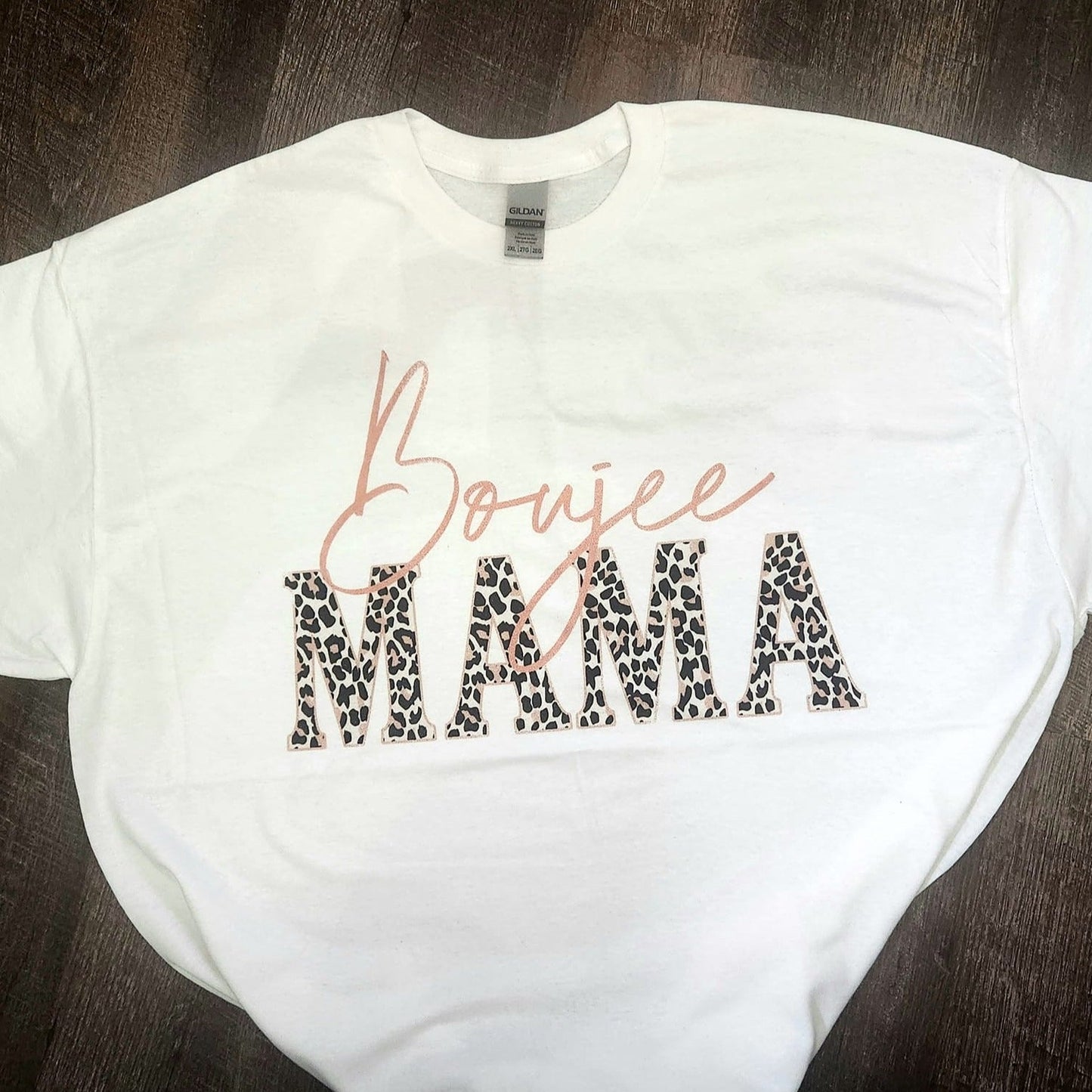 White "Boujee Mama" T-shirt with leopard print lettering displayed on a dark wood background.