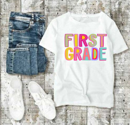 Youth-size First Grade T-shirt in pink with colorful lettering, displayed with jeans and sneakers on a rustic wood background.