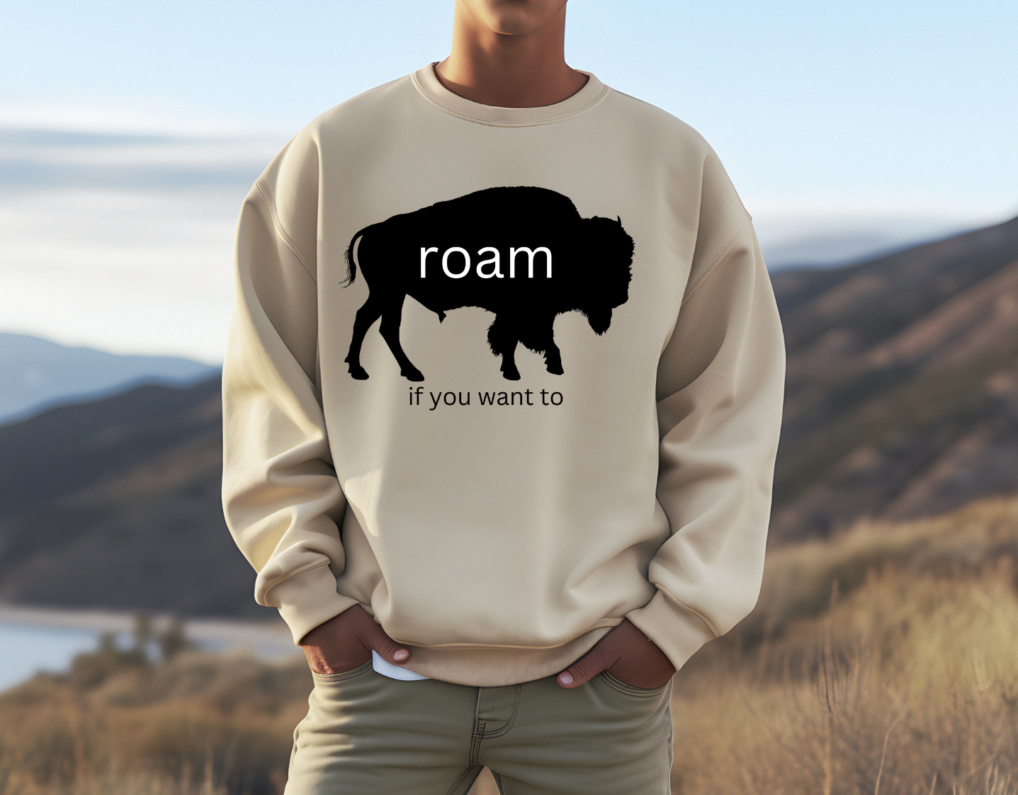 Man wearing "Roam If You Want To" sweatshirt with buffalo design in scenic landscape.