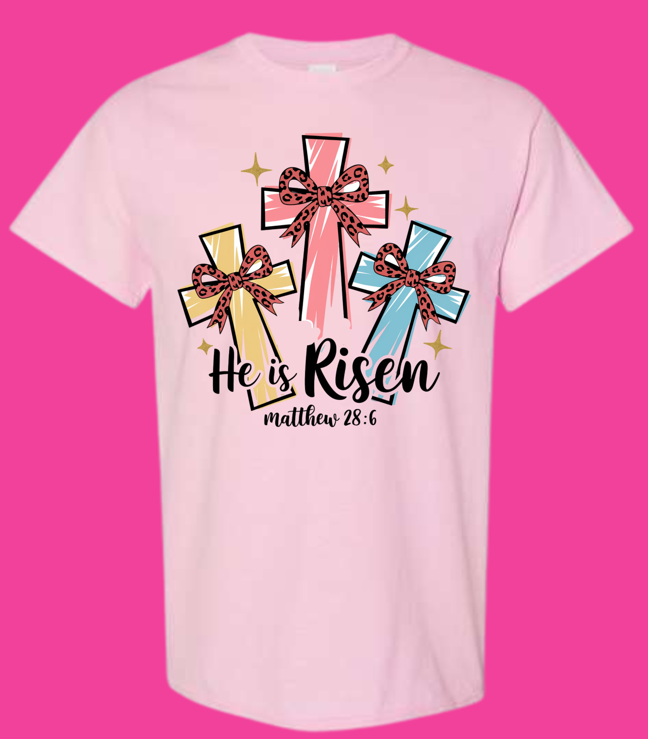 Pink "He Is Risen" T-shirt featuring colorful crosses with bows and the text "Matthew 28:6," celebrating Easter with playful design.