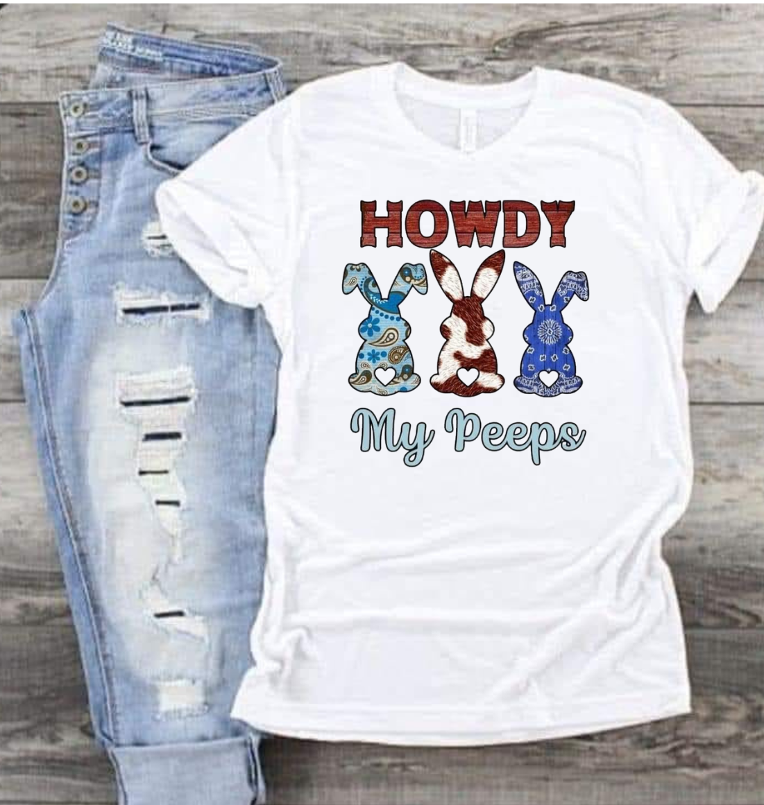 Howdy My Peeps shirt with playful bunny graphic design and text, laid beside ripped jeans.