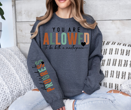 Sweatshirt with "You Are Allowed To Be Both a Masterpiece and Work In Progress" text design, Gildan brand, cotton blend.