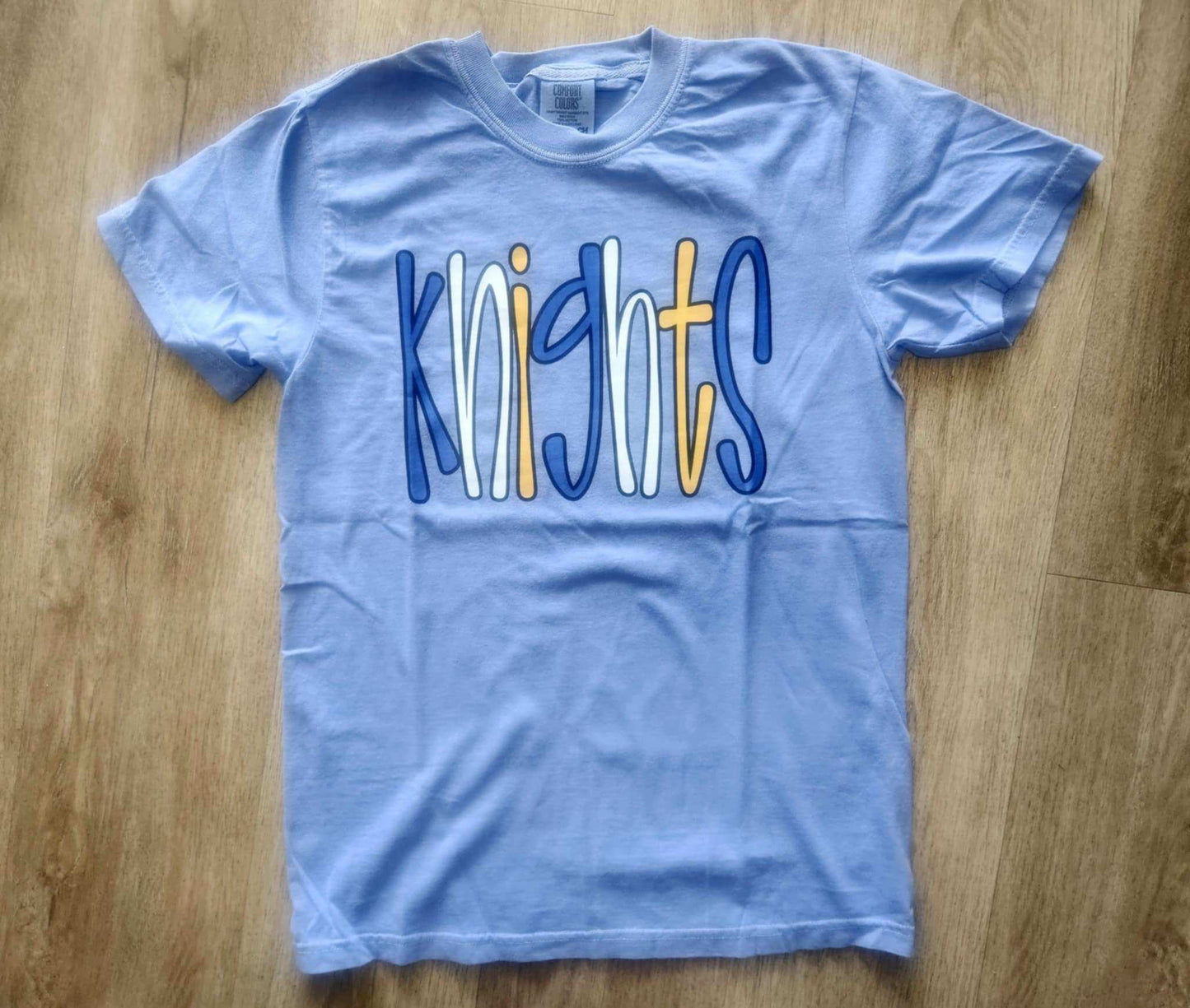 Light blue Knights Bubble Mascot T-shirt with colorful playful lettering on wooden floor background