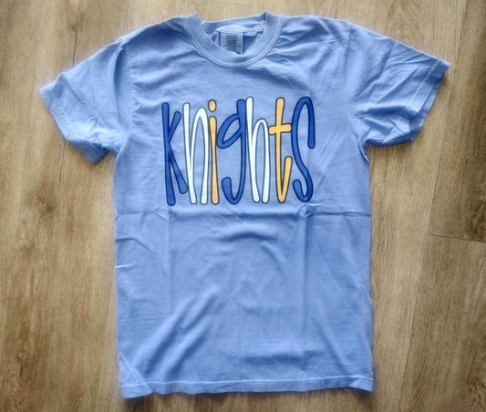 Light blue Knights Bubble Mascot T-shirt with colorful playful lettering on wooden floor background