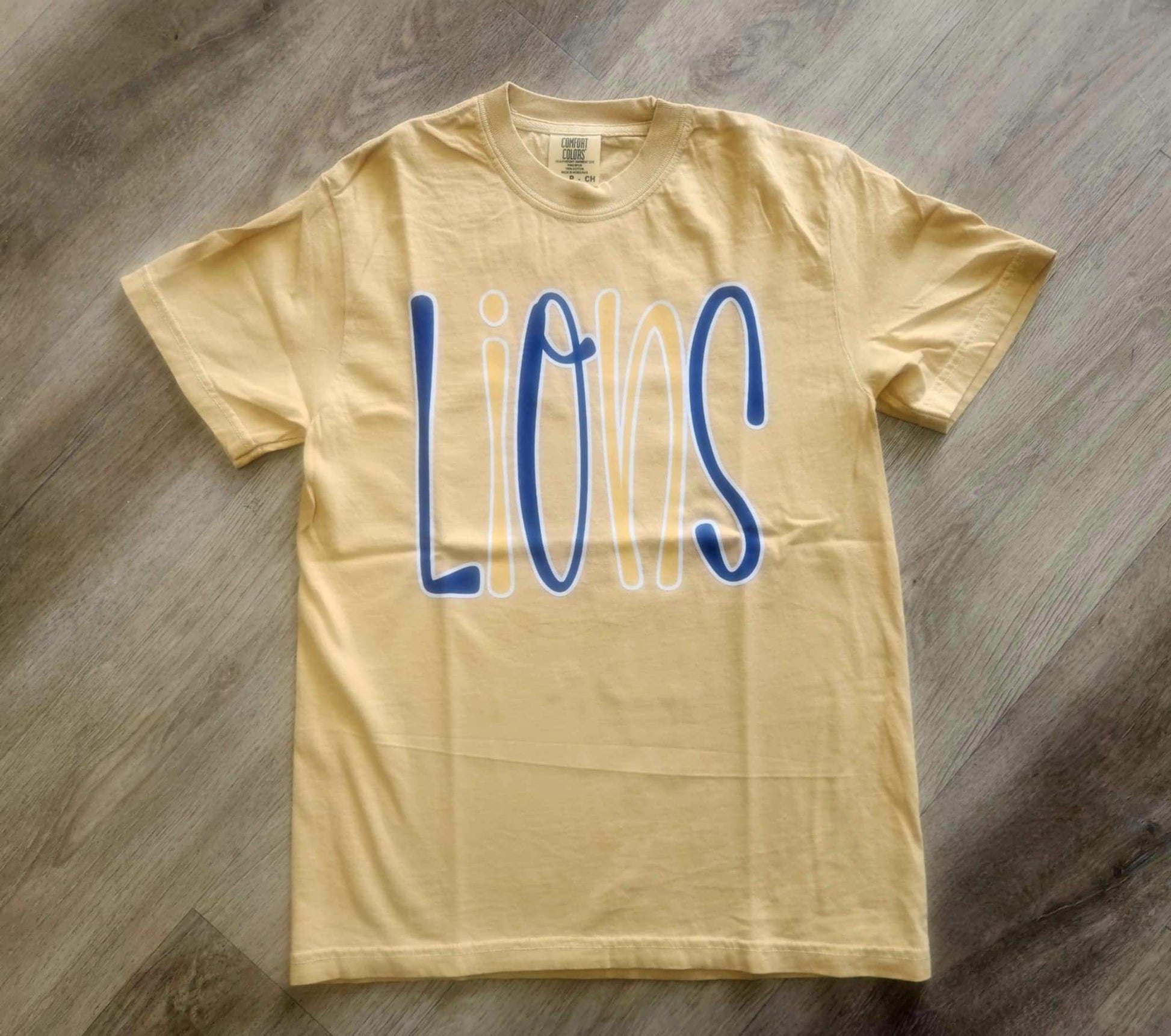 Yellow Lions Bubble Mascot T-shirt with blue and white playful lettering on wooden floor background