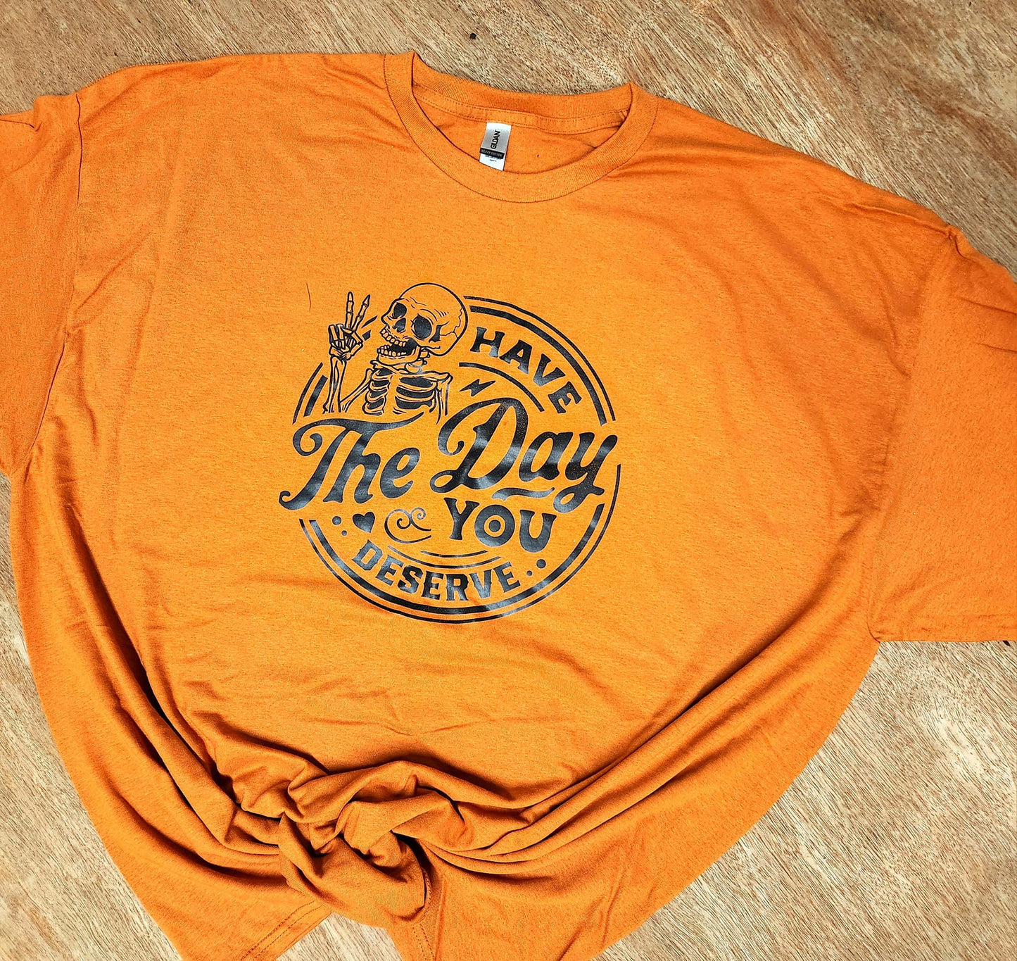 Orange "Have the Day You Deserve" skeleton graphic tee on a wooden surface.