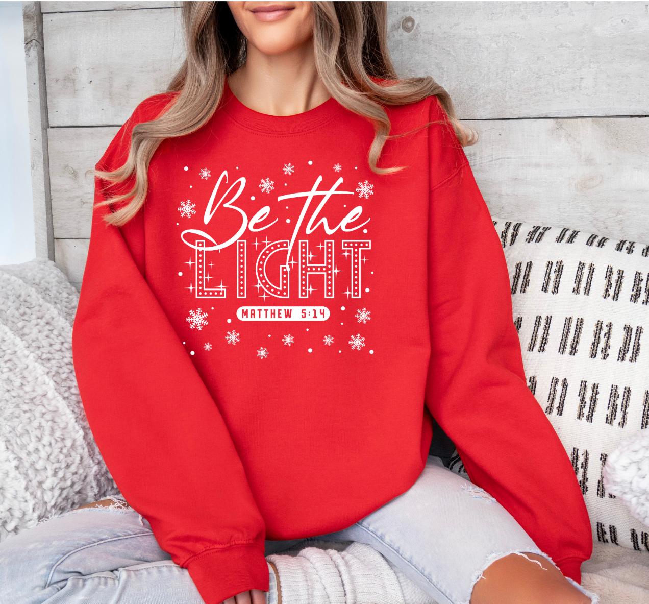 Red 'Be The Light' sweatshirt with white graphic design against a wooden background.
