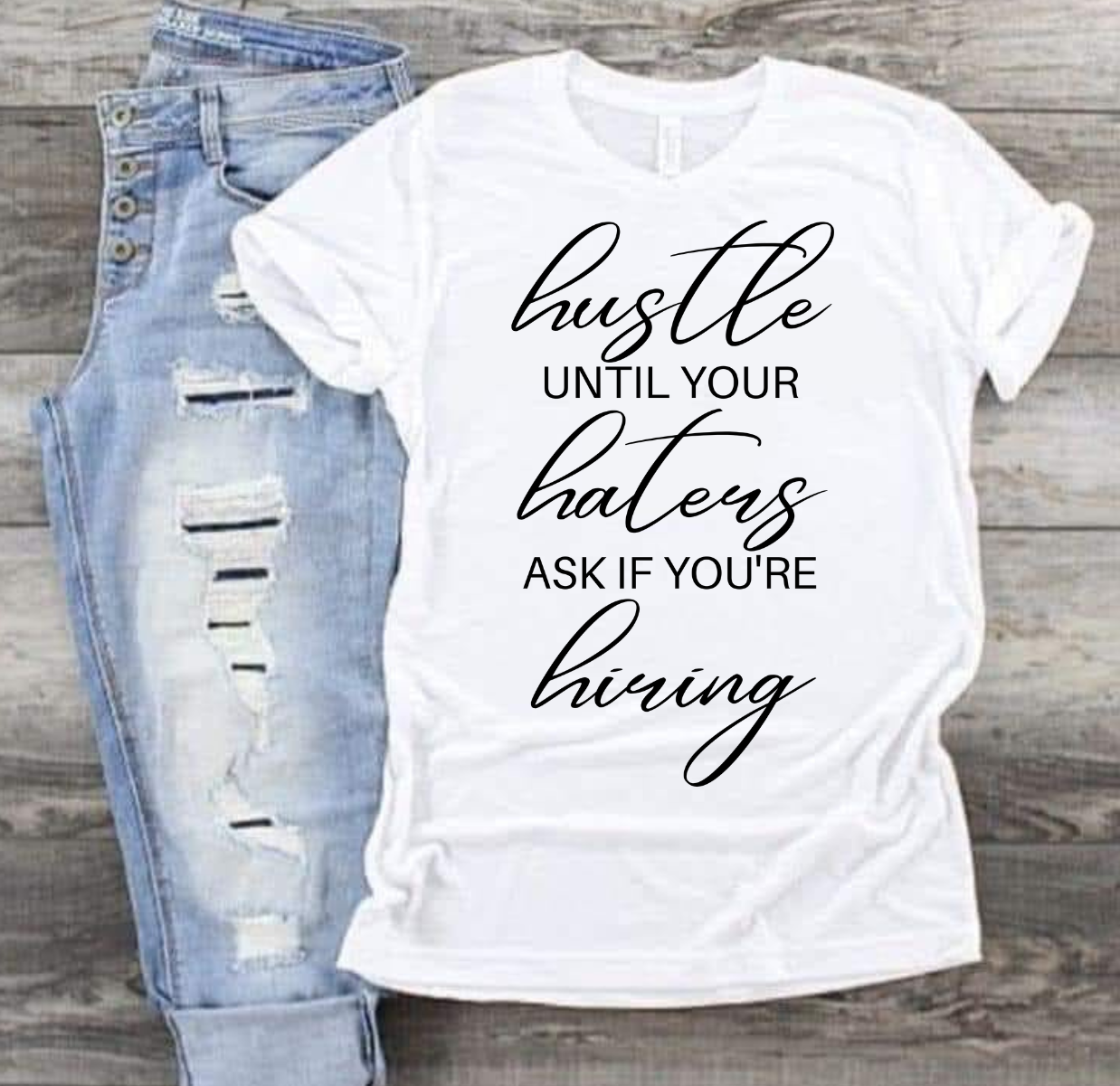 Hustle Til Your Haters T-shirt, bold statement design, next to ripped jeans on wood background.