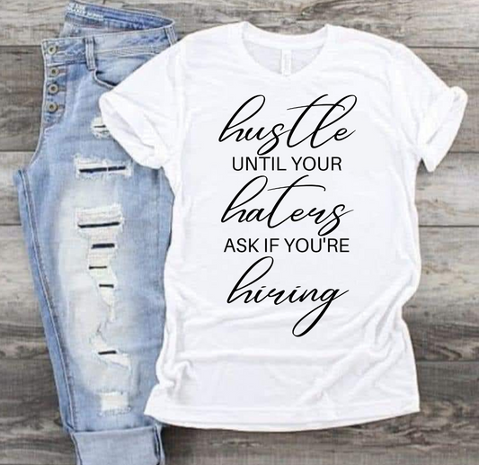 Hustle Til Your Haters T-shirt, bold statement design, next to ripped jeans on wood background.